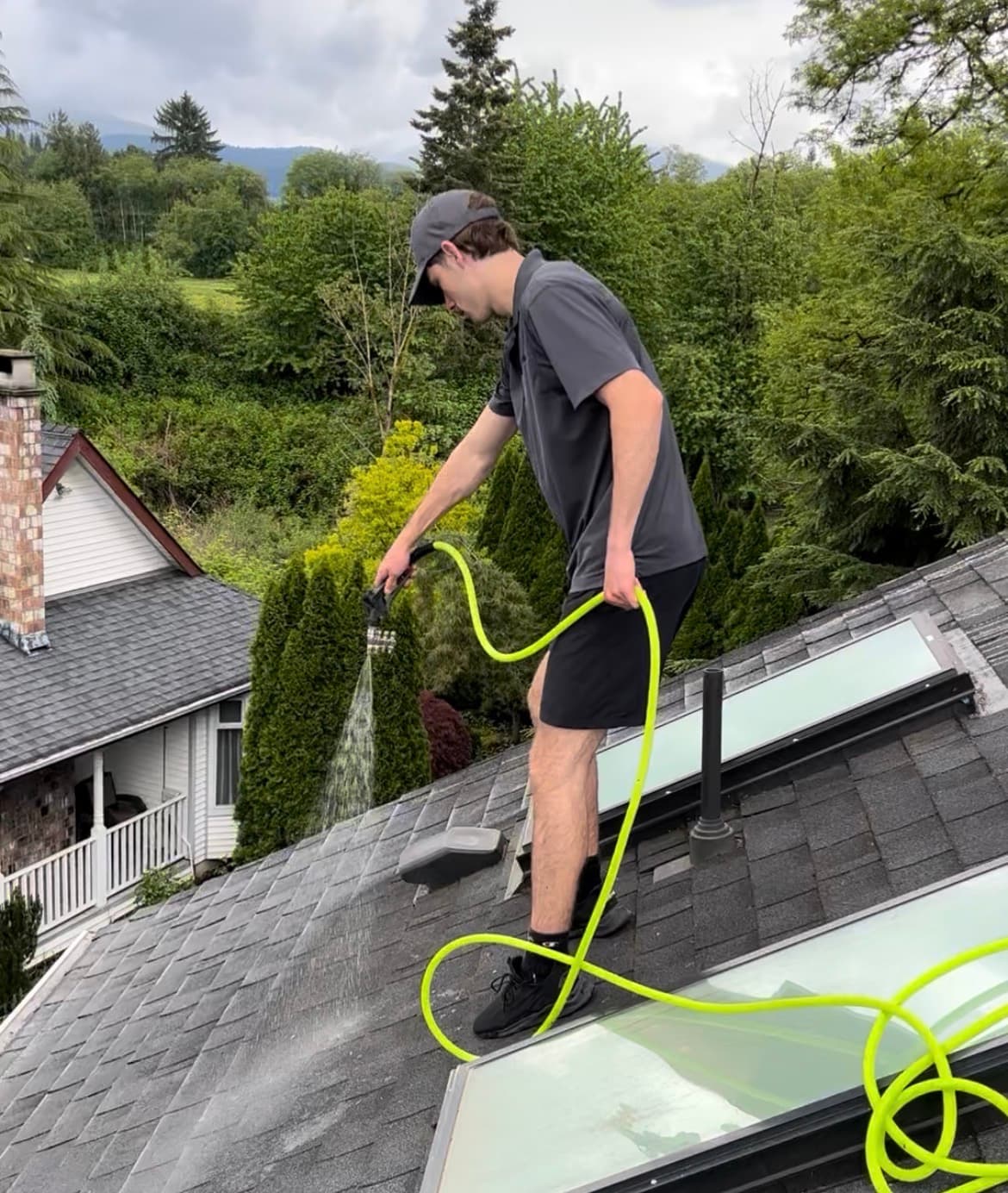 Man on a shingle roof spraying water with a neon green hose near a skylight.